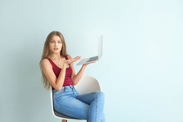 Surprised young woman with laptop sitting on chair against color background - Photo, Image
