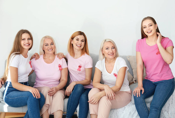 schöne Frauen unterschiedlichen Alters mit rosa Bändern, die auf dem Sofa im Zimmer sitzen. Brustkrebs-Konzept - Foto, Bild