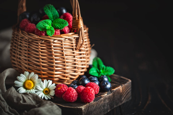 Raspberries and blueberries in a basket with chamomile and leaves on a dark background. Summer and healthy food concept. Background with copy space. Selective focus. - Photo, Image