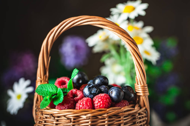 Raspberries and blueberries in a basket with chamomile and leaves on a dark background. Summer and healthy food concept. Selective focus. - Foto, Imagem