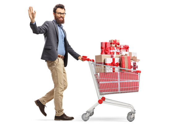 Foto completa de un hombre barbudo saludando y caminando con un carrito de compras lleno de regalos aislados sobre fondo blanco
 - Foto, Imagen