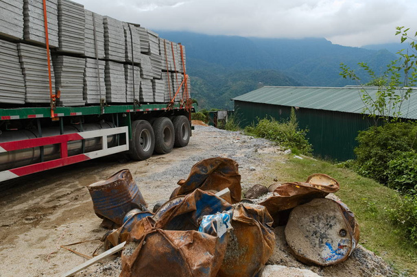 コンクリートブロックを持つ大型貨物トラックは、ゴミのきちんとしたヒープを維持します。建設現場で錆びた損傷した樽の山。背景に雲を持つ山脈。サパ, ベトナム - 写真・画像
