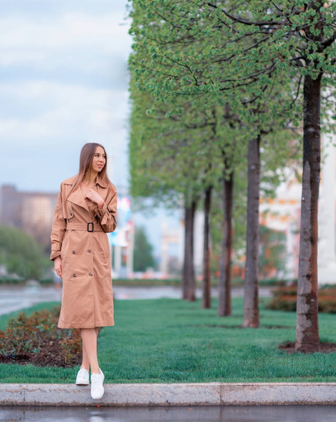 Portrait of a beautiful young brown-haired woman in a brown coat and white shoes in the street in full growth - 写真・画像