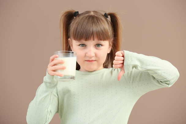 Cute Little Girl z kieliszkiem mleka pokazano kciuka w dół na kolor tła - Zdjęcie, obraz