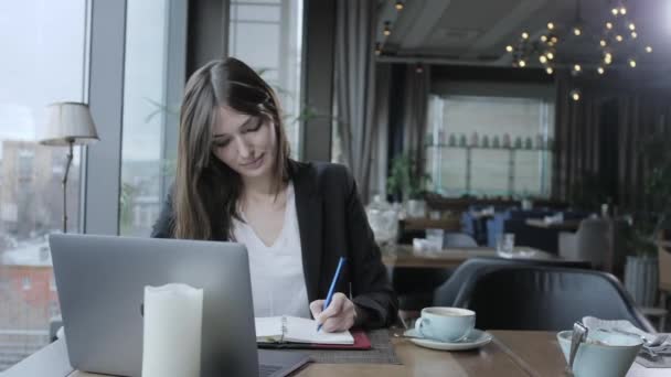 mooie vrouw maken van notities in een notebook. Jonge zakenvrouw zittend in coffeeshop aan houten tafel. Op tafel is grijs aluminium laptop. Schema's en maakt belangrijke opmerkingen - Video