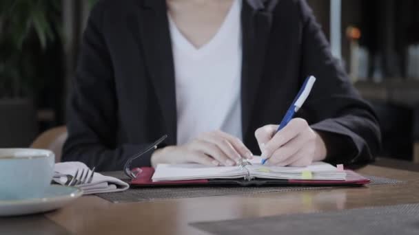 Κοντινό χέρι μιας γυναίκας που κάνει σημειώσεις σε ένα σημειωματάριο. Νεαρή επιχειρηματίας που κάθεται στην καφετέρια στο ξύλινο τραπέζι. Προγραμματίζει και κάνει σημαντικές σημειώσεις - Πλάνα, βίντεο