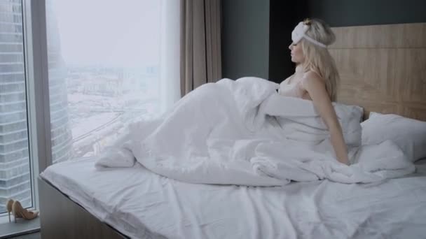 Dobré ráno v hotelovém pokoji. Mladá žena sedí na pohodlné posteli v masce na spaní na hlavě. Okno mrakodrapu na pozadí. - Záběry, video