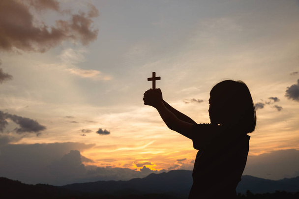 Το χέρι ενός κοριτσιού που κρατά ένα σταυρό κατά την Ανατολή, την ευχαριστία θεραπεία ευλόγησε τον Θεό βοηθώντας μετανοήσει Καθολικό Πάσχα. Προσευχή προς τον Θεό χριστιανική θρησκεία έννοια φόντο. - Φωτογραφία, εικόνα