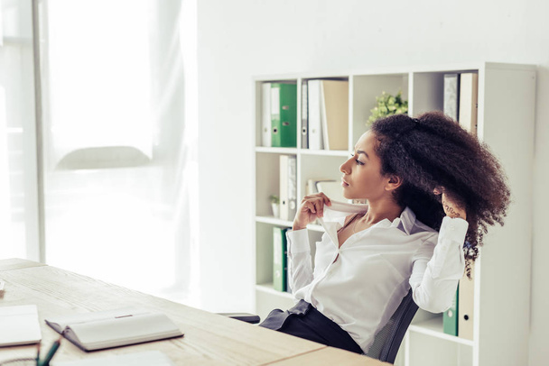 hübsche afrikanisch-amerikanische Geschäftsfrau schaut weg, während sie am Arbeitsplatz sitzt und im Büro unter Hitze leidet - Foto, Bild
