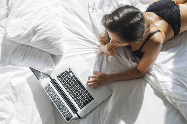Sexy woman in black lingerie lying on the bed and using laptop - Fotografie, Obrázek