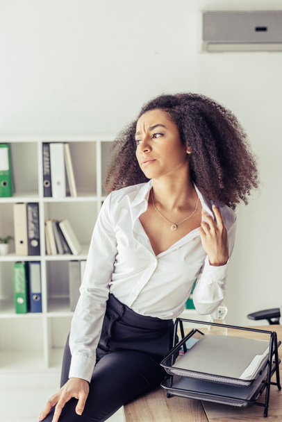 exhausted african american businesswoman sitting on desk while suffering from heat in office - 写真・画像