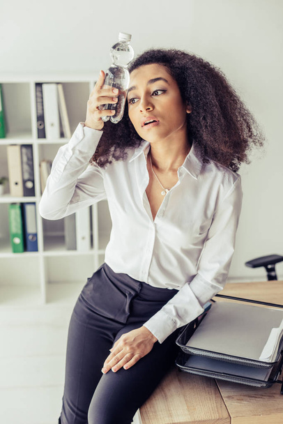 mujer de negocios afroamericana agotada sosteniendo botella de plástico con agua cerca de la cabeza mientras sufre de calor en la oficina
 - Foto, Imagen