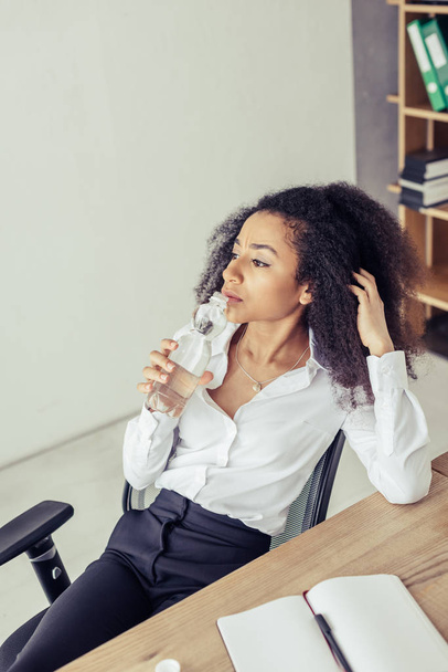 mulher de negócios afro-americana pensativa bebendo água e olhando para longe enquanto sofre de calor no escritório
 - Foto, Imagem