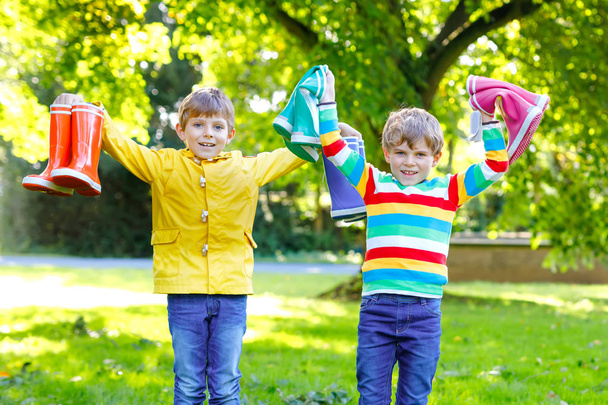 2人の小さな男の子、カラフルな雨のブーツがたくさんあるかわいい兄弟。異なるゴムブーツやジャケットの子供。雨の秋のための履物。健康的な双子と親友が屋外で楽しんでいます - 写真・画像