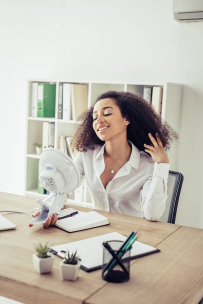 cheerful african american businesswoman holding electric fun while suffering from heat in office - Photo, image
