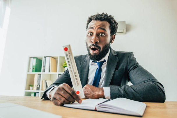 järkyttynyt afrikkalainen amerikkalainen liikemies katselee lämpömittari kärsiessään lämpöä toimistossa
 - Valokuva, kuva