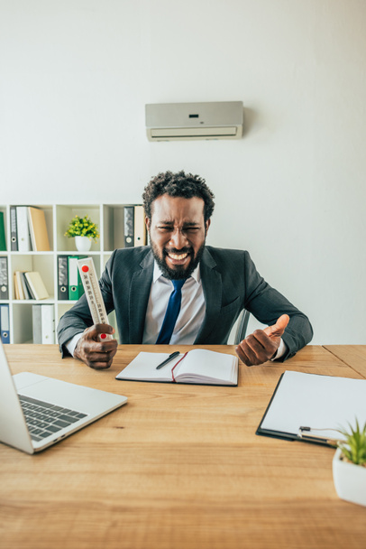 irritated african american businessman holding thermometer and gesturing while suffering from summer heat in office - Photo, Image