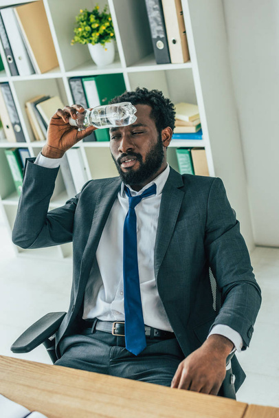 unhappy african american businessman holding plastic bottle near head while suffering from summer heat in office - Photo, Image