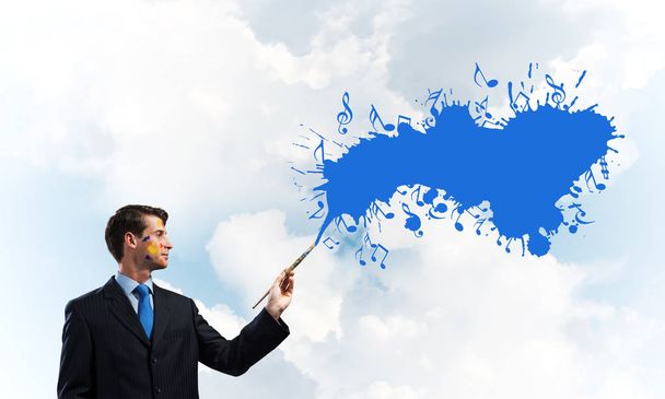 Uomo d'affari di successo in abito nero gesticolando e sorridendo mentre in piedi con spruzzi musicali di colore blu contro cielo nuvoloso vista sullo sfondo
. - Foto, immagini
