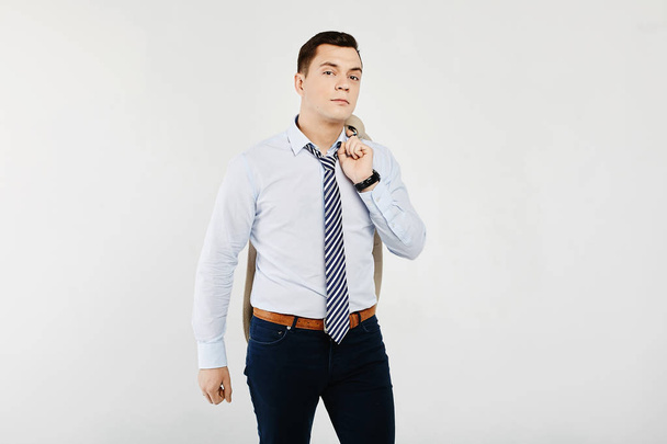 Young and stylish businessman in the white shirt with a striped tie and in the dark jeans holding a beige blazer on the shoulder, isolated at white background - Photo, image