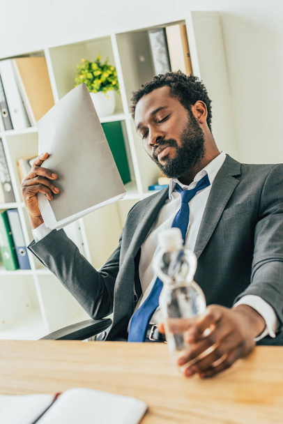 オフィスで熱に苦しんでいる間、水とフォルダのボトルを保持している疲れたアフリカ系アメリカ人のビジネスマン - 写真・画像