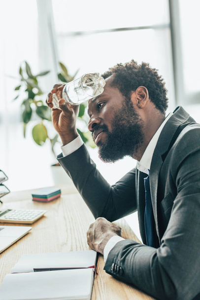 bearded african american businessman holding plastic bottle with water while sitting at workplace - 写真・画像