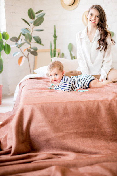 Beautiful young mother with long dark hair in homely comfortable clothes plays with her young son in a cozy bedroom with live green plants and furniture made from natural materials - Φωτογραφία, εικόνα
