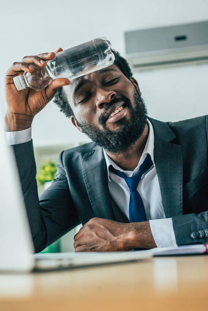 focalizzazione selettiva di un uomo d'affari afroamericano esausto che tiene una bottiglia con acqua vicino alla testa mentre soffre di calore in ufficio
  - Foto, immagini