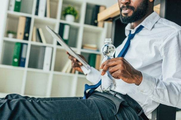 vista cortada do empresário americano africano segurando garrafa de água e acenando com pasta enquanto sentado no chão no escritório
 - Foto, Imagem