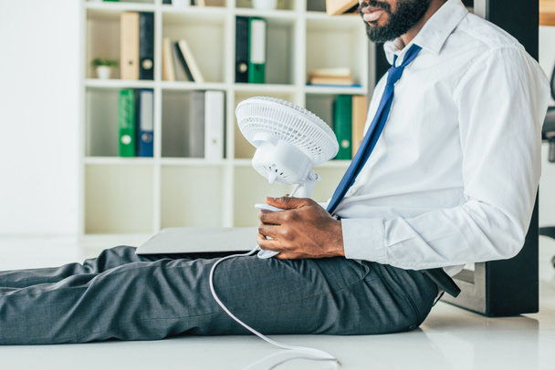 vista ritagliata di uomo d'affari afro-americano seduto sul pavimento e che tiene ventilatore elettrico mentre soffre di calore in ufficio
 - Foto, immagini