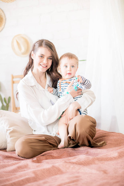 Beautiful young mother with long dark hair in homely comfortable clothes plays with her young son in a cozy bedroom with live green plants and furniture made from natural materials - Фото, изображение