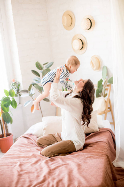 Beautiful young mother with long dark hair in homely comfortable clothes plays with her young son in a cozy bedroom with live green plants and furniture made from natural materials - Foto, immagini
