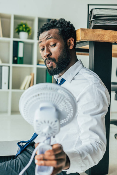 guapo afroamericano hombre de negocios sosteniendo soplado ventilador eléctrico mientras está sentado en el suelo en la oficina
 - Foto, imagen
