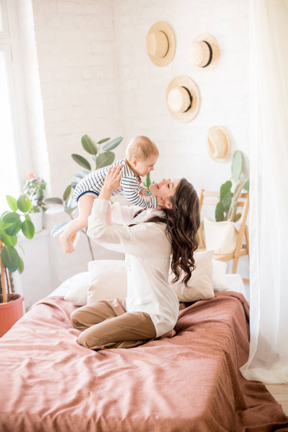 Beautiful young mother with long dark hair in homely comfortable clothes plays with her young son in a cozy bedroom with live green plants and furniture made from natural materials - Fotoğraf, Görsel