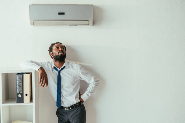 young african american businessman standing under air conditioner and holding hand on hip - Foto, Imagen