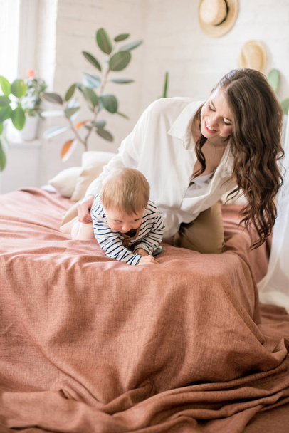 Beautiful young mother with long dark hair in homely comfortable clothes plays with her young son in a cozy bedroom with live green plants and furniture made from natural materials - Foto, Imagen
