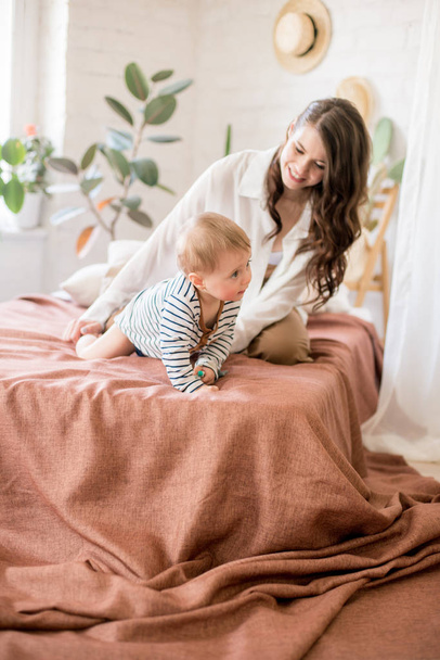 Beautiful young mother with long dark hair in homely comfortable clothes plays with her young son in a cozy bedroom with live green plants and furniture made from natural materials - Foto, immagini