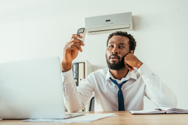 homme d'affaires afro-américain tenant climatiseur télécommande tout en souffrant de chaleur au bureau
 - Photo, image
