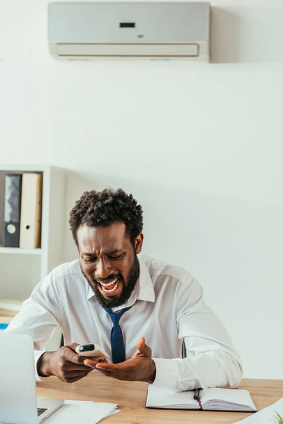 раздраженный африканский американский бизнесмен, глядя на пульт дистанционного управления кондиционером, страдающий от жары в офисе
 - Фото, изображение
