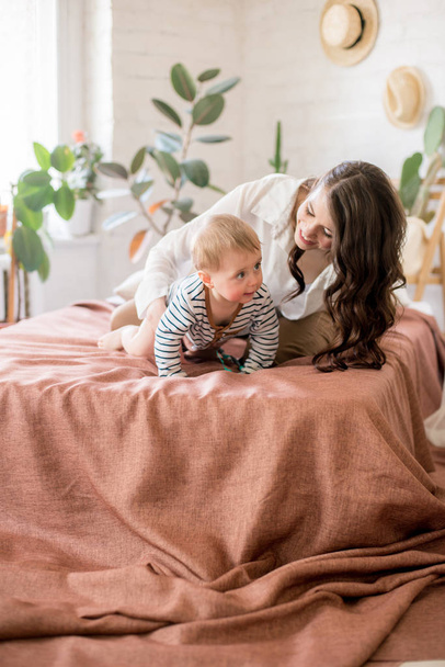 Beautiful young mother with long dark hair in homely comfortable clothes plays with her young son in a cozy bedroom with live green plants and furniture made from natural materials - Valokuva, kuva