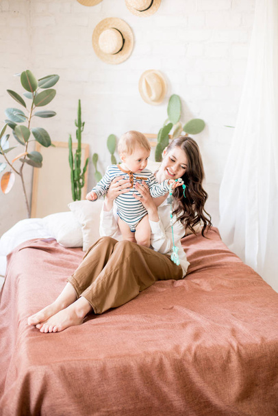 Beautiful young mother with long dark hair in homely comfortable clothes plays with her young son in a cozy bedroom with live green plants and furniture made from natural materials - Photo, Image