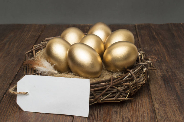 Nest with golden eggs with a tag and place for text on a wooden background. The concept of successful retirement - 写真・画像