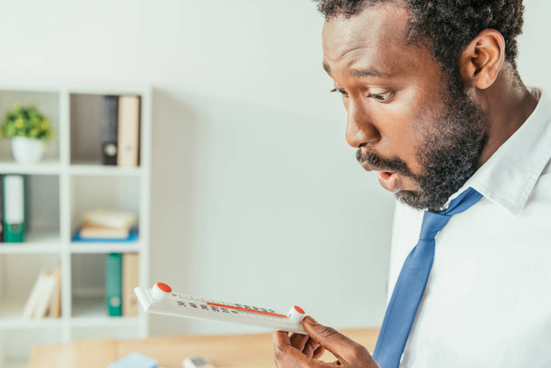 shocked african american businessman looking at thermometer showing high temperature in office - Photo, Image