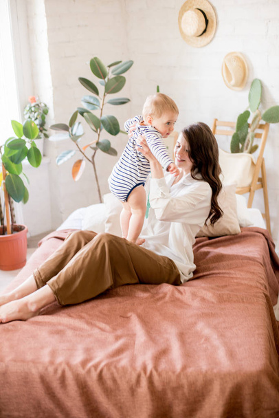 Beautiful young mother with long dark hair in homely comfortable clothes plays with her young son in a cozy bedroom with live green plants and furniture made from natural materials - Foto, afbeelding