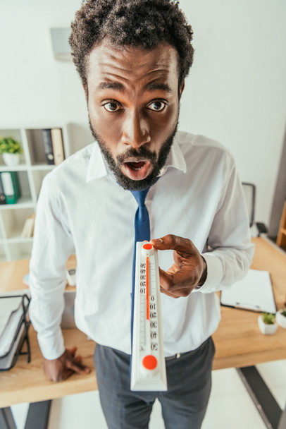 shocked african american businessman looking at camera and showing thermometer  - Zdjęcie, obraz