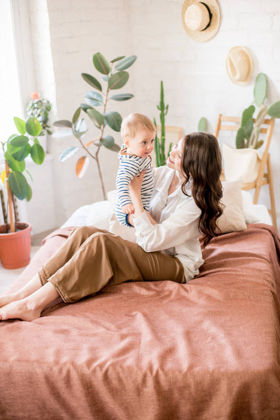 schöne junge Mutter mit langen dunklen Haaren in heimeliger bequemer Kleidung spielt mit ihrem kleinen Sohn in einem gemütlichen Schlafzimmer mit lebendigen grünen Pflanzen und Möbeln aus natürlichen Materialien - Foto, Bild