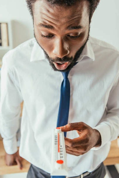 шокированный африканский американский бизнесмен смотрит на термометр, показывающий высокую температуру в офисе
 - Фото, изображение