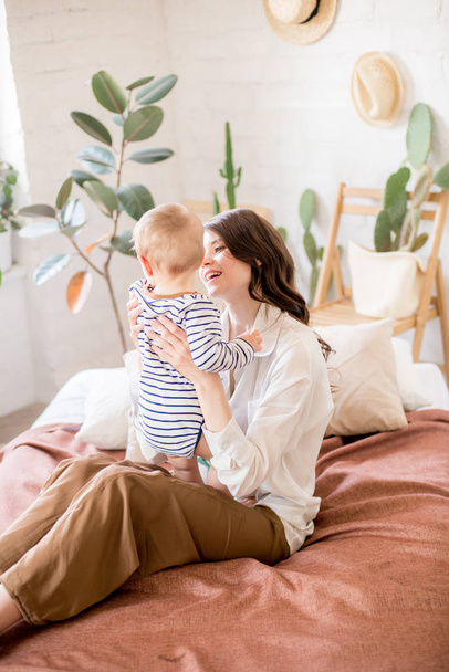 Beautiful young mother with long dark hair in homely comfortable clothes plays with her young son in a cozy bedroom with live green plants and furniture made from natural materials - Foto, imagen