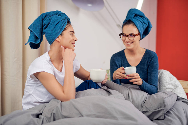 Teo όμορφη Καυκάσιος φίλες με πετσέτες στο κεφάλι κάθεται στο κρεβάτι στο υπνοδωμάτιο, πίνοντας καφέ και κουτσομπολεύοντας. - Φωτογραφία, εικόνα