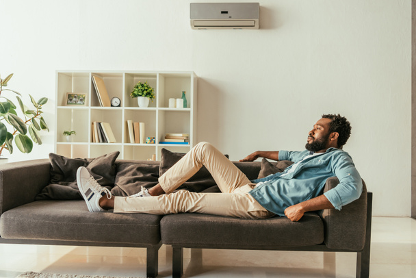 joven afroamericano hombre acostado en sofá en casa mientras que sufre de calor de verano
 - Foto, Imagen